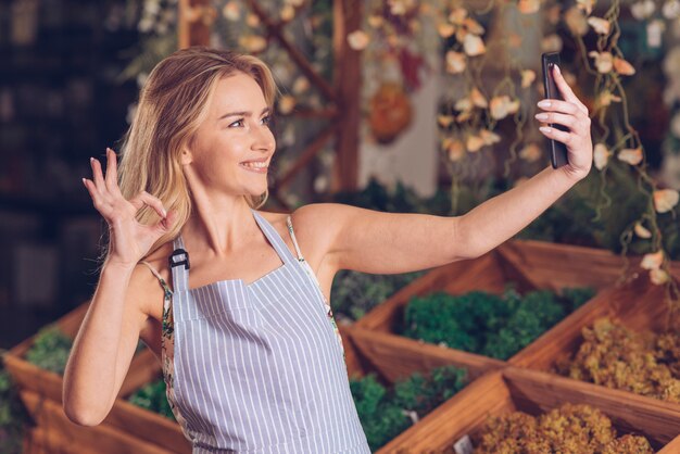 Florista de sexo femenino joven sonriente que toma el selfie en el teléfono elegante que muestra gesto aceptable en la tienda