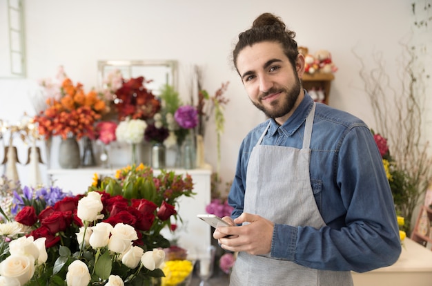Florista de sexo masculino joven feliz que sostiene el teléfono móvil en la mano que mira a la cámara