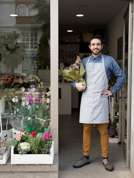 Florista de sexo masculino joven sonriente confiado que se coloca en la entrada de la floristería que sostiene el ramo disponible