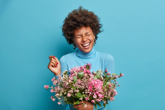 Floristería alegre posa con un hermoso ramo de flores, se ríe alegremente, ha cerrado los ojos, viste poloneck azul, posa en interiores