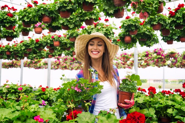 Floristería hermosa mujer atractiva con sombrero caminando a través de invernadero sosteniendo flores en macetas y control de plantas