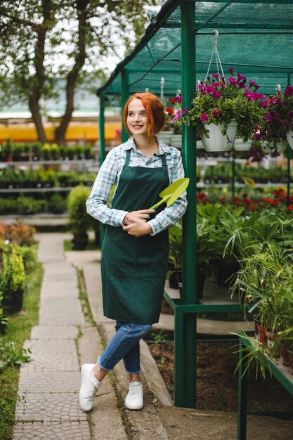 Floristería sonriente en delantal de pie con una pequeña pala de jardín en la mano. Señorita mirando felizmente a un lado mientras trabaja con flores