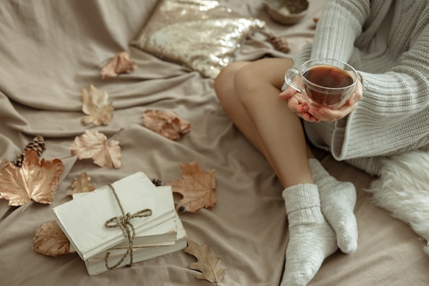 Fondo de otoño acogedor con piernas femeninas en calcetines calientes, una taza de té y hojas de otoño en la cama.