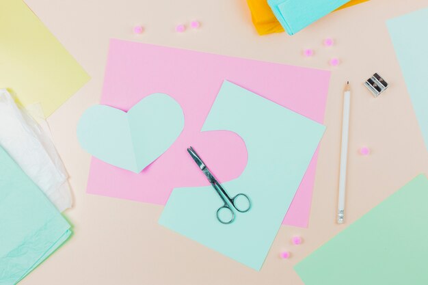 Forma de corazón papel azul con tijera; Lápiz y sacapuntas sobre fondo beige.