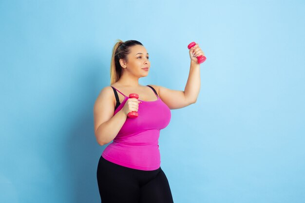 Formación de jóvenes caucásicos plus size modelo femenino sobre fondo azul. Concepto de deporte, emociones humanas, expresión, estilo de vida saludable, cuerpo positivo, igualdad. Entrenando con las pesas, copyspace.