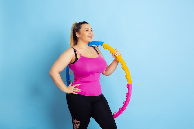 Formación de jóvenes caucásicos plus size modelo femenino sobre fondo azul. Concepto de deporte, emociones humanas, expresión, estilo de vida saludable, cuerpo positivo, igualdad. Haciendo ejercicio, posando con el aro.