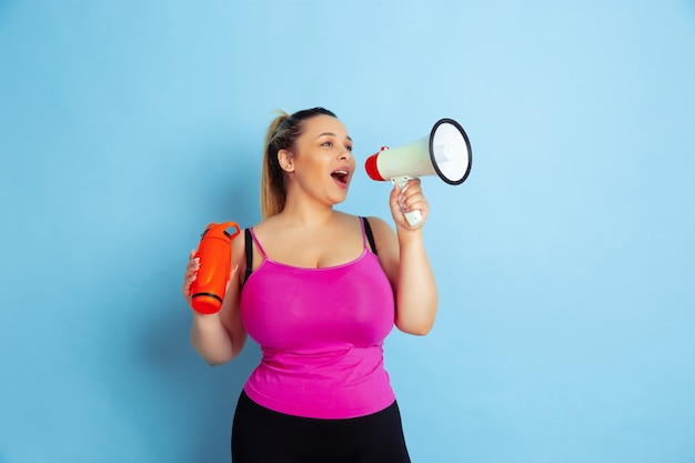 Formación del modelo femenino caucásico joven del tamaño extra grande en fondo azul. Concepto de deporte, emociones humanas, expresión, estilo de vida saludable, cuerpo positivo, igualdad. Posando con botella y bocapaz.