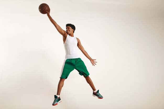 Foto de acción de un joven atleta negro feliz con camisa blanca y pantalones cortos verdes saltando alto para agarrar una pelota de baloncesto vintage en blanco