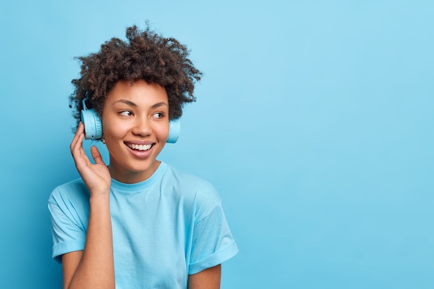 Foto de una adolescente complacida con cabello afro escucha música a través de auriculares inalámbricos de una lista de reproducción vestida con ropa casual aislada sobre un espacio de copia de pared azul para su anuncio