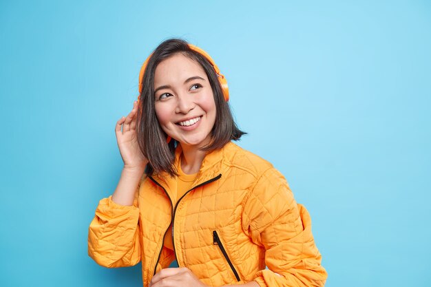 La foto de una adolescente guapa con apariencia oriental escucha música en auriculares modernos sonríe ampliamente viste una chaqueta naranja de moda aislada sobre una pared azul. Gran lista de reproducción. Amante de la música