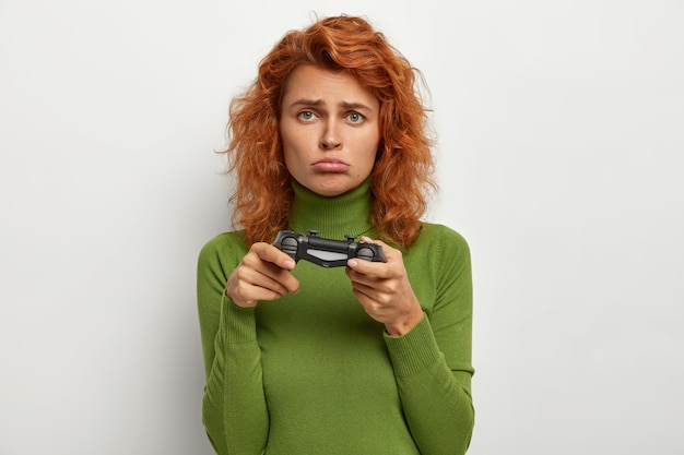 Foto de una adolescente pelirroja que juega con el joystick, tiene una expresión infeliz, pierde el videojuego, pasa el tiempo libre en casa, es un verdadero jugador. Personas, ocio, concepto de entretenimiento.