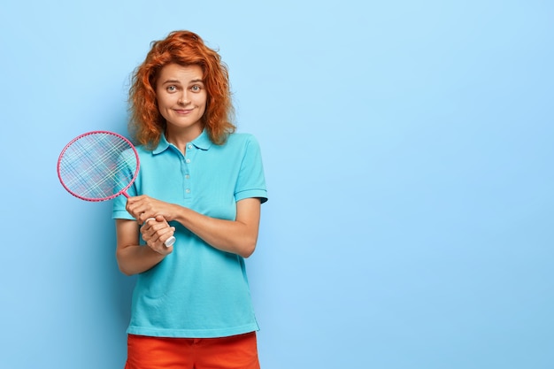 Foto de adolescente pelirrojo se encuentra con raqueta de bádminton, espera a que su compañero juegue un juego deportivo, vestido con ropa casual