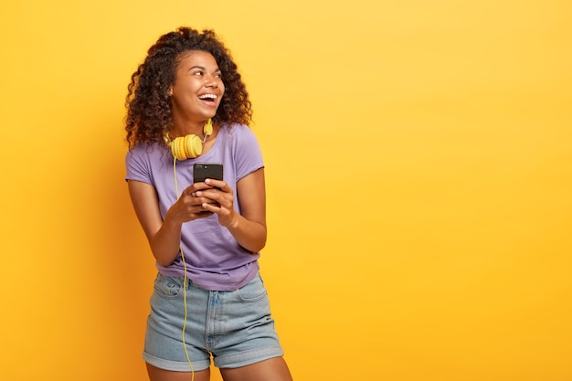 Foto de una adolescente sonriente con corte de pelo afro, usa un teléfono inteligente para escuchar música en la lista de reproducción, usa auriculares, mira positivamente a un lado