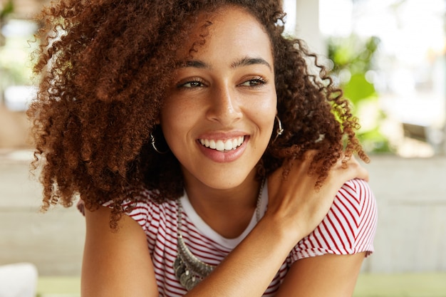 Foto de adorable hermosa mujer sonriente con peinado afro, vestida con camiseta a rayas, tiene expresión positiva y soñadora, piensa en algo agradable. Mujer joven con amplia sonrisa brillante