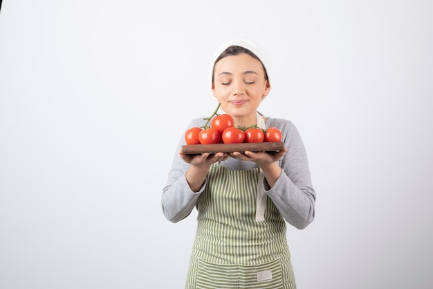 Foto de adorable joven huele tomates rojos sobre pared blanca