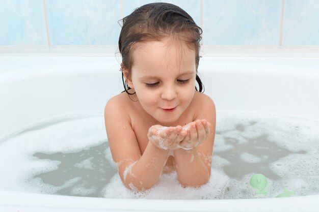 Foto de adorable niña con cabello mojado jugando con espuma de jabón en la bañera