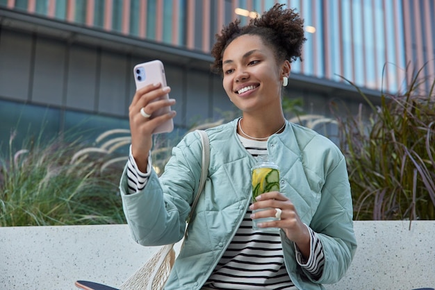 Foto gratuita una foto al aire libre de una adolescente feliz con dos bollos rizados se toma una selfie a través de un teléfono inteligente bebe una bebida de desintoxicación usa un suéter casual a rayas y una chaqueta lleva una bolsa de red pasa el tiempo libre en un entorno urbano