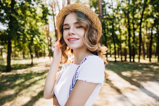 Foto al aire libre de chica espectacular escalofriante en el hermoso bosque. Señora alegre con sombrero de paja vintage posando en el parque.