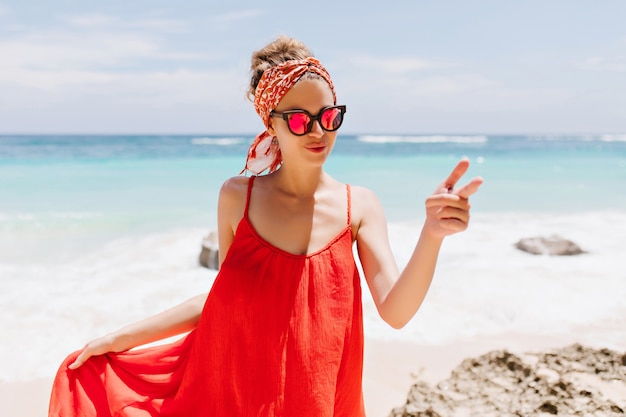 Foto al aire libre de elegante niña caucásica lleva gafas brillantes durante el descanso cerca del océano. Retrato de hermosa mujer bronceada en traje rojo escalofriante en la playa salvaje.