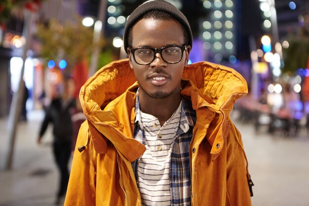 Foto al aire libre de un elegante turista de piel oscura con abrigo de invierno, sombrero y gafas caminando por la ciudad de noche, haciendo autostop por Europa, de pie en medio de la calle y mirando con una sonrisa