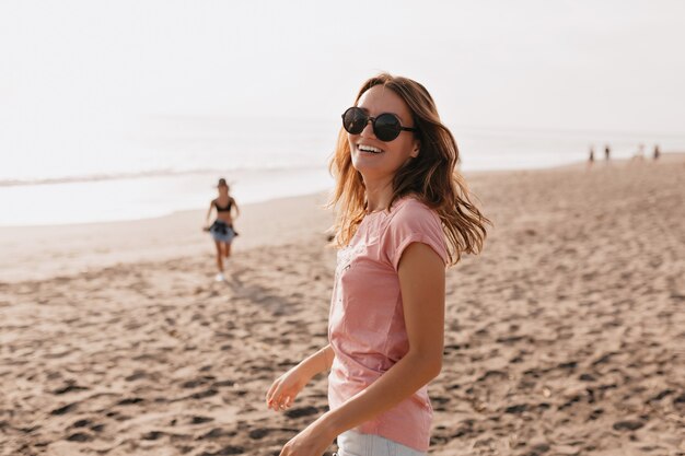 Foto al aire libre de feliz modelo de mujer joven en camiseta de verano de pie contra el cielo azul y la playa de arena Mujer divirtiéndose en un día de verano