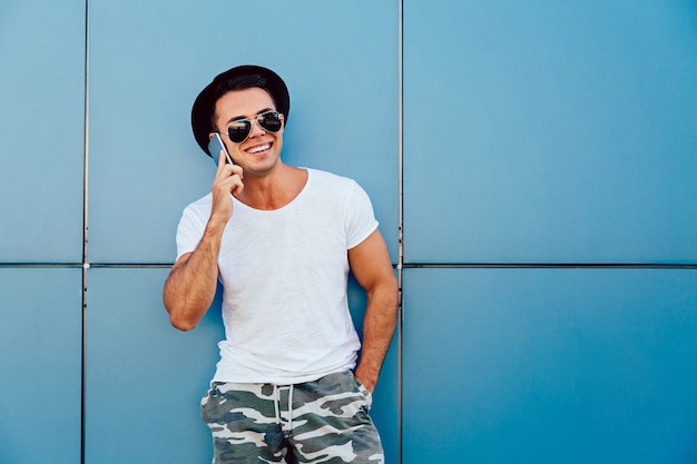 Foto al aire libre de hombre joven con estilo alegre llamando con teléfono inteligente, llevaba sombrero negro