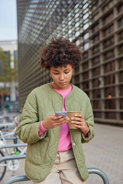 Una foto al aire libre de una joven de cabello rizado bebe café aromático y navega por Internet en un teléfono móvil usa una chaqueta mira videos en línea pasa tiempo libre en poses de la ciudad contra un fondo borroso