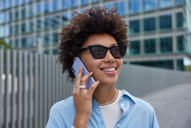 Una foto al aire libre de una joven morena rizada tiene una conversación telefónica con gafas de sol y una camisa azul casual sonríe alegremente habla en llamadas digitales a un amigo con un fondo borroso