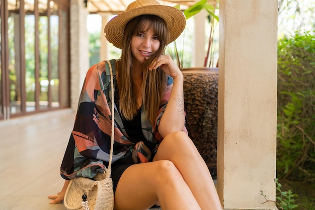 Foto al aire libre de una magnífica mujer caucásica con sombrero de paja y elegante atuendo de verano disfrutando de vacaciones tropicales en un complejo moderno