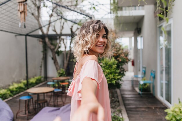 Foto al aire libre de la maravillosa chica rubia mirando por encima del hombro. Mujer europea de pelo corto inspirada en traje rosa que se coloca cerca del restaurante de la calle.