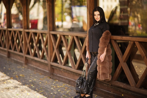 Foto al aire libre de moda de mujer sensual hermosa con cabello oscuro en ropa elegante y lujoso abrigo de piel contra terraza de madera en la ciudad de otoño