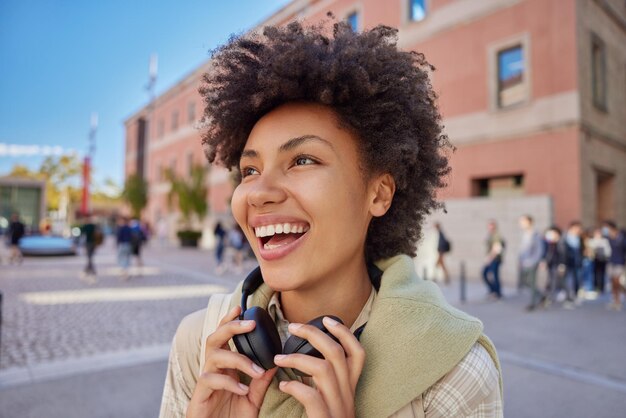 Una foto al aire libre de una mujer alegre con el pelo rizado vestido con ropa informal que usa auriculares estéreo alrededor del cuello tiene una expresión alegre camina en una calle concurrida contra un fondo borroso Concepto de estilo de vida