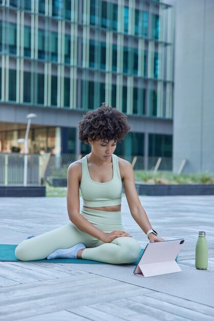 La foto al aire libre de una mujer deportiva de cabello rizado que va a realizar cambios de clase de fitness en línea en la cámara en una tableta digital tiene entrenamiento regular en prácticas al aire libre, aprendizaje de yoga. Deporte en lugar de urabn