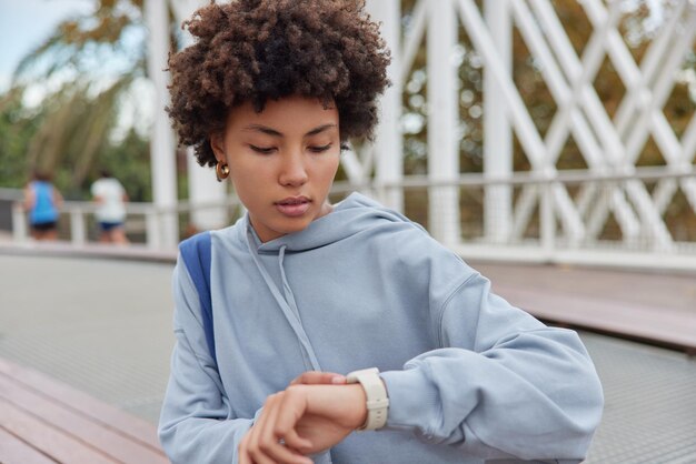 Una foto al aire libre de una mujer milenaria seria con el pelo rizado revisa el tiempo en el reloj de pulsera esperando a alguien vestido con poses de capucha azul contra un fondo borroso durante el día Concepto de estilo de vida