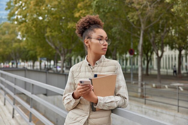 Una foto al aire libre de una mujer pensativa de cabello rizado usa un teléfono móvil para chatear en línea sostiene poses de cuadernos en la ciudad contra el parque usa anteojos redondos y el chaleco tiene una expresión pensativa Estilo de vida