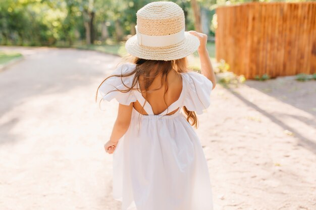 Foto al aire libre de la parte posterior de la niña con pie de piel bronceada en la calle en una mañana soleada. Encantadora niña lleva sombrero de paja decorado con cinta y vestido blanco bailando en el parque.