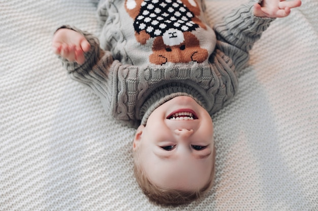 Foto al revés de un niño adorable en suéter caliente acostado en la cama y riendo