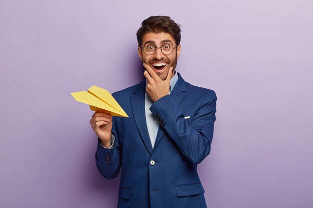 Foto de alegre empresario próspero es dueño de una gran empresa, sonríe positivamente, usa gafas y traje transparentes, lanza un avión de papel