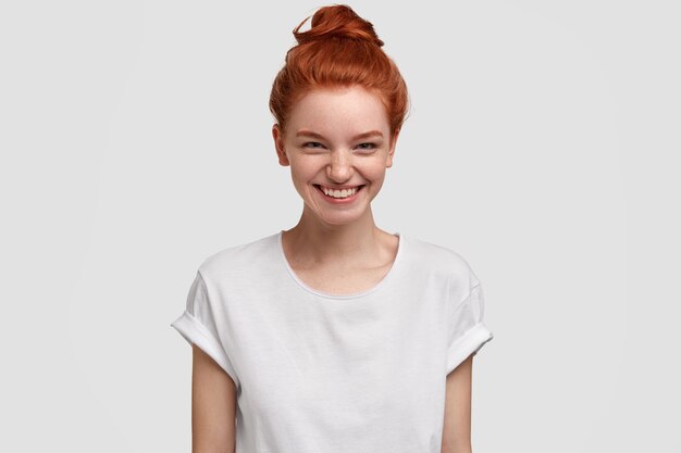 Foto de alegre encantadora adolescente pelirroja sonríe con expresión curiosa e interesada, acepta oferta maravillosa, viste camiseta blanca informal, modelos de interior