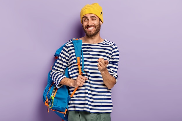 Foto de alegre hombre sonriente que el viajero le señala con el dedo índice, lleva una mochila, viste un sombrero amarillo y un suéter con rayas, expresa su elección, lo recoge, aislado en una pared púrpura