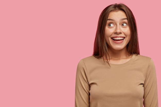 Foto de alegre joven europea tiene una amplia sonrisa, se ve positiva y con expresión soñadora a un lado, vestida con un suéter informal, aislada sobre una pared rosa con espacio de copia a la izquierda