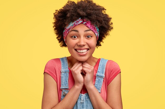 Foto de alegre mujer afroamericana mantiene las manos juntas cerca de la barbilla, sonríe ampliamente