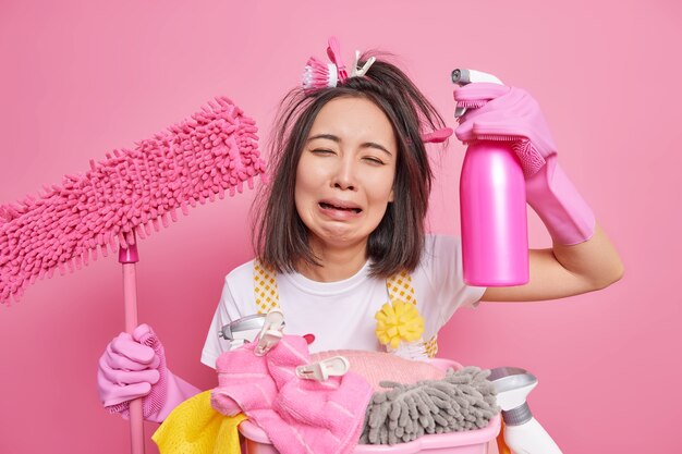 Foto de ama de casa estresada llora de desesperación por estar harta de la limpieza de la casa sostiene la fregona y el detergente tiene expresión triste funciona sobre la casa aislada sobre fondo rosa. Limpiador de fatiga.