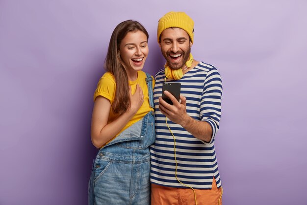 Foto de amigos alegres que pasan tiempo libre juntos, miran algo en el teléfono inteligente, están de pie cerca uno del otro