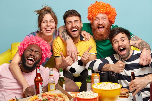 Foto de amistosos compañeros que se abrazan y sonríen felices, animan con el equipo favorito ganador, pasan un buen rato juntos viendo un emocionante partido de fútbol, beben cerveza y comen comida rápida. Soporte de fans divertidos