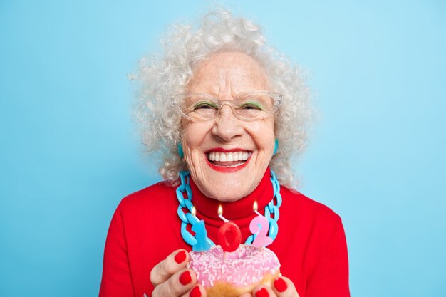 Foto de una anciana de moda alegre sonríe con dientes sostiene un dulce donut glaseado con velas encendidas hace un deseo en su cumpleaños usa un suéter rojo, vasos transapantes posa en el interior