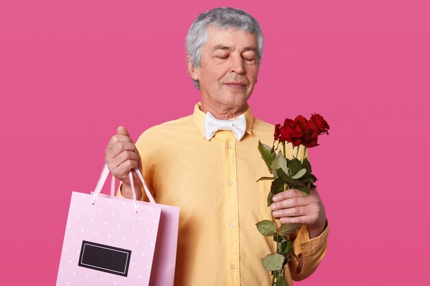 Foto de apuesto hombre maduro, viste elegante camisa amarilla, corbatín, sostiene un ramo de rosas rojas y una bolsa rosa con regalo para su esposa