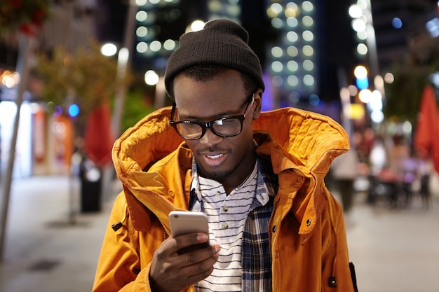 Foto de un apuesto joven estudiante afroamericano que camina por la ciudad de noche, sostiene el teléfono móvil, usa wifi urbano, navega por las imágenes en las redes sociales. Tecnología moderna y comunicación