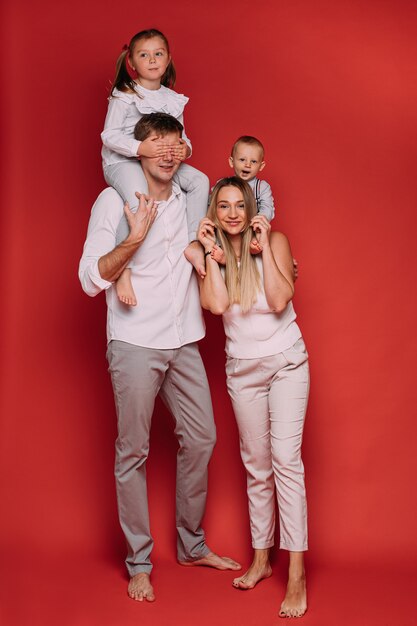 Foto de archivo de cuerpo entero de amoroso padre y madre con hijos sobre los hombros posando sobre fondo rojo. Hija cerrando los ojos del padre con las manos.