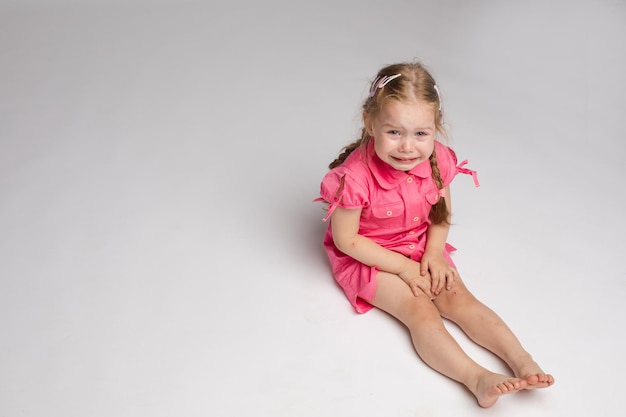 Foto de archivo de una dulce niña con trenzas en vestido rosa llorando mientras se sienta en el suelo con los pies descalzos Ella está mirando a la cámara mientras solloza